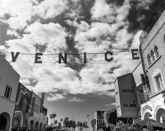May include: Black and white photo of a street in Venice, California. The street is lined with buildings and palm trees. A sign that reads "VENICE" is strung across the street.
