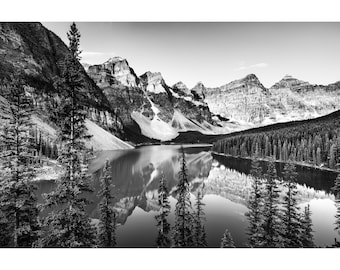 May include: A black and white photograph of a mountain lake with snow-capped mountains in the background. The lake is surrounded by trees and reflects the mountains in the water.