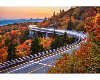 May include: A winding road with a concrete bridge over a valley. The trees are in vibrant fall colors, with shades of red, orange, yellow, and green. The road is empty and the sky is a soft pink and orange.