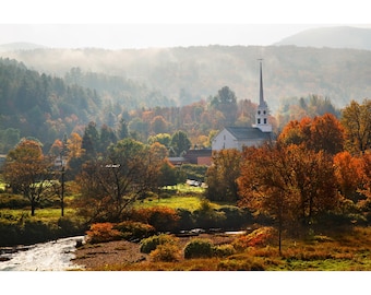May include: A scenic autumn landscape featuring a white church with a tall steeple, surrounded by trees with vibrant orange and red foliage. A river flows through the foreground, with a backdrop of rolling hills and a misty sky.