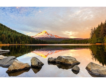 May include: A scenic landscape photograph of a mountain reflected in a calm lake. The mountain peak is covered in snow and illuminated by the setting sun. The foreground features rocks, and the surrounding area is filled with evergreen trees.