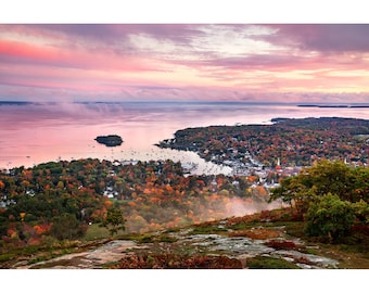 May include: A panoramic view of a coastal town with a harbor filled with boats. The town is surrounded by trees with fall foliage, and the sky is a vibrant pink and orange.