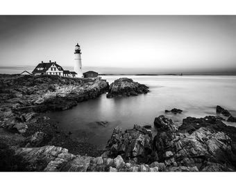 May include: A black and white landscape photograph featuring a tall, cylindrical lighthouse standing on a rocky coastline. A multi-story building is attached to the base of the lighthouse, overlooking a calm, expansive body of water.