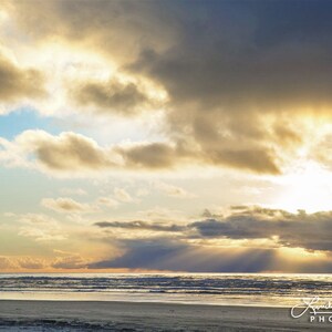 May include: A scenic photograph of a beach at sunset. The sky is filled with dramatic clouds in shades of gray and gold, with the sun breaking through, casting rays of light over the ocean. The beach is visible in the foreground.