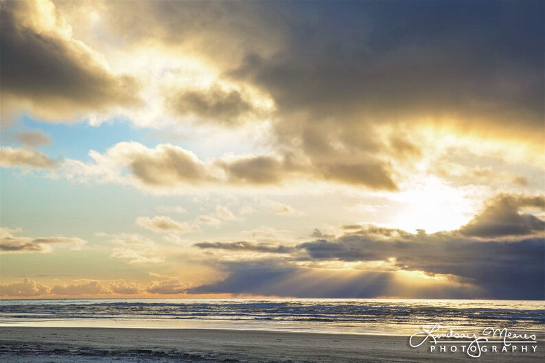 May include: A scenic photograph of a beach at sunset. The sky is filled with dramatic clouds in shades of gray and gold, with the sun breaking through, casting rays of light over the ocean. The beach is visible in the foreground.