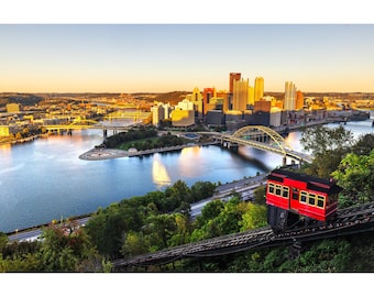 May include: A scenic view of Pittsburgh, Pennsylvania, featuring a red funicular car ascending a hillside. The city skyline is visible in the background, with bridges spanning the river. The sky is a gradient of blue and orange.