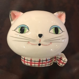 May include: A white ceramic cat figurine with a smiling face, green eyes, and a black nose. The cat wears a red and black plaid scarf. The ears have pink inner details. The figurine is a decorative item.