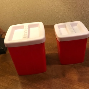 May include: Two red plastic containers with white lids. The lids have the words "Sugar" and "Milk" printed on them.