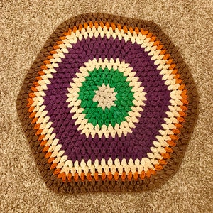 May include: A crocheted rug with a hexagonal shape. The rug has a brown, orange, cream, purple, and green color pattern.