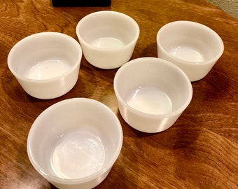 May include: Five white ceramic ramekins, also known as custard cups, are arranged on a wooden surface. The ramekins are all empty and have a smooth, glossy finish.