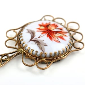 May include: A vintage hand mirror with a floral design. The mirror has a white ceramic face with an orange and red flower and grey leaves. The ornate gold-coloured metal frame has a decorative handle.