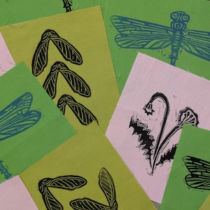 May include: A collection of handmade art prints featuring nature-inspired linocut designs. The prints showcase dragonflies, seed pods, and floral motifs in black ink on green and pink paper. The prints are arranged in a layered composition.