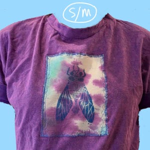 May include: Purple t-shirt with a tie-dye design featuring a blue insect graphic. The shirt has a crew neck and short sleeves. The size tag says "S/M".