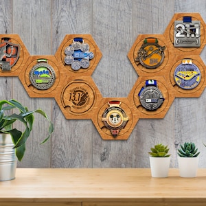 Puede incluir: Expositor de medallas hexagonal de madera con ocho soportes para medallas. El expositor está montado en una pared de madera gris claro. Los soportes para medallas están dispuestos en forma de panal. Hay varias medallas expuestas en los soportes, incluida una medalla de Mickey Mouse.