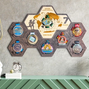 Puede incluir: Un expositor hexagonal de madera con siete compartimentos para medallas. El expositor presenta un mapa del mundo con el texto "Seven Continents" y "Mark Fuller" en la parte superior. El expositor está montado en una pared gris.