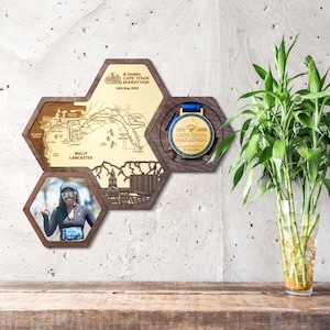 May include: A wall-mounted wooden display featuring three connected hexagonal frames. One frame holds a map of the Sanlam Cape Town Marathon with the text Mally Lancaster, another displays a gold marathon medal, and the third holds a photograph.