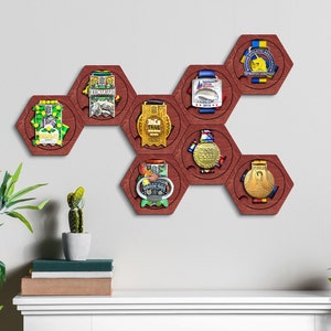 Puede incluir: Un expositor de medallas hexagonal de madera con siete soportes para medallas. Cada soporte es un hexágono con un marco de madera rojo y un inserto de plástico transparente para mostrar las medallas. El expositor está montado en una pared blanca.