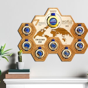 Puede incluir: Un mapa del mundo de madera con siete pantallas hexagonales para medallas. Cada hexágono presenta una medalla de oro con una cinta azul y el contorno de un continente. El texto "World Marathon Challenge 2020" se muestra en el mapa. El texto "Jenn Solomon #34" se muestra debajo del mapa.