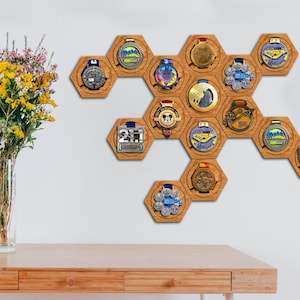 Puede incluir: Un expositor hexagonal de madera con 14 soportes para medallas. El expositor está montado en una pared blanca. Las medallas son de varios colores y diseños. Un jarrón de flores amarillas y blancas está sobre una mesa de madera en primer plano.