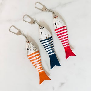 May include: Three wooden fish keychains with striped patterns. The fish are painted white with red, blue, and orange stripes. The keychains are attached to silver key rings.
