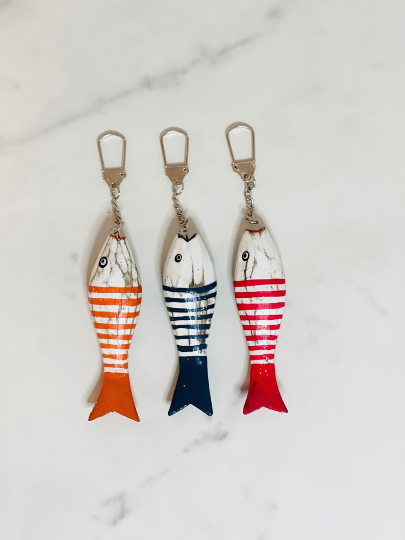 May include: Three wooden fish keyrings with striped designs. The fish are painted white with red, orange, and blue stripes. Each keyring has a silver metal ring and chain.