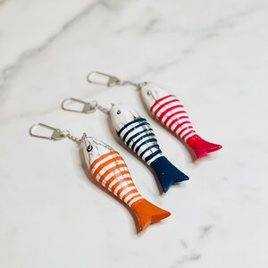 May include: Three wooden fish keychains with striped patterns. The fish are painted white with red, blue, and orange stripes. The keychains are attached to silver chains.