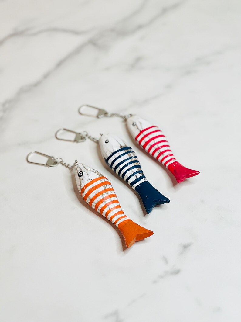 May include: Three wooden fish keychains with striped patterns. The fish are painted white with red, blue, and orange stripes. The keychains are attached to silver chains.