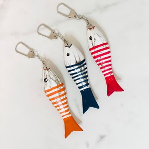 May include: Three wooden fish keyrings with striped patterns. The fish are painted white with red, blue, and orange stripes. Each fish has a silver keyring attached.