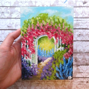 May include: A painting of a white garden archway covered in pink flowers, with a path leading through a field of purple and blue flowers.