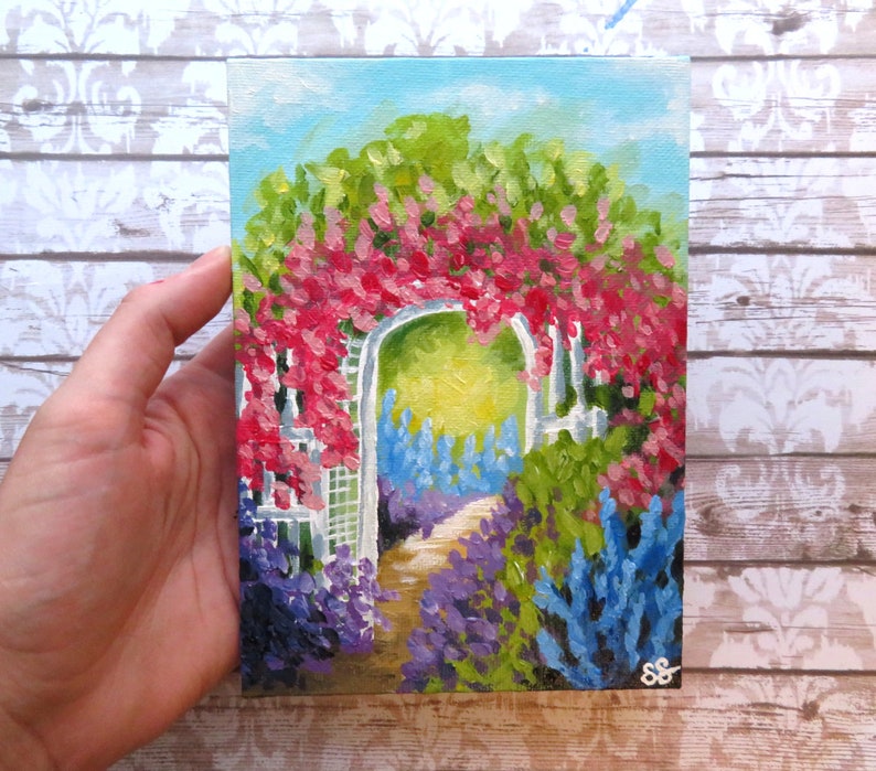 May include: A painting of a white garden archway covered in pink flowers, with a path leading through a field of purple and blue flowers.