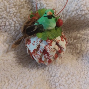 May include: A decorative ornament featuring a green bird with a black mustache perched atop a woven fabric ball. The ball is patterned with red floral designs on a white background. A red loop is attached for hanging.