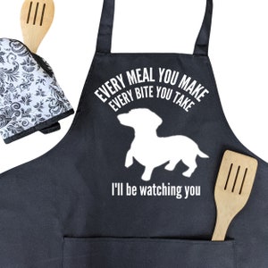 May include: Black kitchen apron with a white silhouette of a dachshund and the text "Every meal you make every bite you take I'll be watching you".