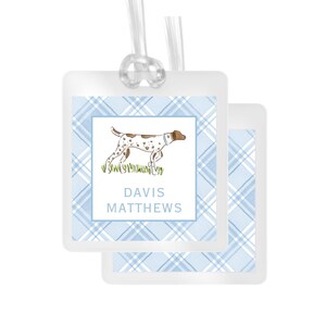 May include: A square luggage tag with a light blue and white plaid pattern. The center features a white square with a brown and white spotted dog standing in green grass. The text DAVIS MATTHEWS is printed in blue below the dog. A clear plastic loop is attached.