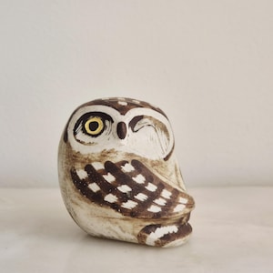 Puede incluir: Figura de búho de cerámica con un ojo guiñado. El búho es principalmente blanquecino con alas y marcas en la cabeza marrones y blancas. El ojo tiene un iris amarillo y un contorno marrón. La figura es un objeto decorativo.