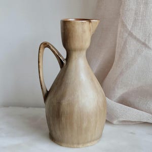 Puede incluir: Una jarra de cerámica beige con cuello delgado y asa curva. La jarra tiene un pico para verter y un cuerpo redondeado. La superficie tiene un aspecto texturizado y moteado con vetas marrones oscuras. La jarra es probablemente para uso decorativo o funcional.