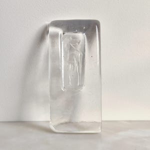 Puede incluir: Un jarrón de cristal transparente, rectangular, con una figura tallada en el centro. El jarrón tiene forma rectangular con bordes redondeados. La figura parece ser una forma humana estilizada. El cristal es transparente.