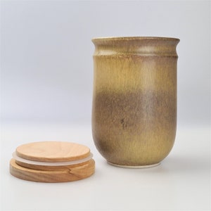 May include: A ceramic jar with a lid. The jar is a light brown color with a darker brown band around the middle. The lid is made of wood and has a light brown color.