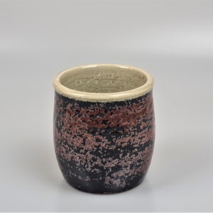 Puede incluir: Vaso de cerámica hecho a mano con superficie texturizada. La taza presenta una base negra con un patrón moteado en tonos rojos y marrones. El borde es de color beige claro.