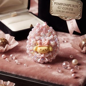 May include: An oval-shaped decorative accessory featuring a central yellow Pompompurin character figure, adorned with pink bows and an array of iridescent pearls and beads. The item is displayed near a black box labeled POMPOMPURIN G7 COVER - LUXURY EDITION.