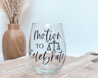 Op de afbeelding: Een helder, steelvrij wijnglas met de woorden "Motion to Celebrate" in zwart schrift en een weegschaal. Het glas staat op een houten plank, met een vaas met droogbloemen op de achtergrond.