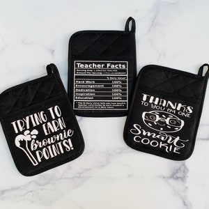 May include: Three black oven mitts with white text. One mitt says "Trying to earn brownie points!" with a cartoon brownie. Another mitt says "Teacher Facts" with a nutrition label listing "Hard Work", "Encouragement", "Dedication", "Inspiration", and "Education" all at 100% daily value. The third mitt says "Thanks to you I'm one smart cookie" with a cartoon cookie wearing glasses.