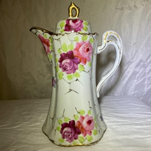 May include: White porcelain coffee pot with a lid, featuring a floral design of pink and purple roses with green leaves. The pot has a gold trim and handle, and a crackle pattern.