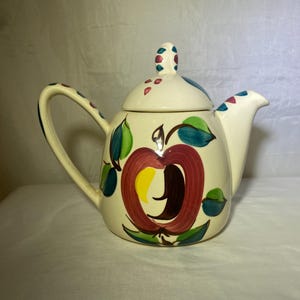 Puede incluir: Tetera de cerámica color crema con tapa y asa. La tetera está decorada con un gran diseño de manzana roja con hojas verdes y un centro amarillo. El asa y la tapa están adornadas con puntos de colores.