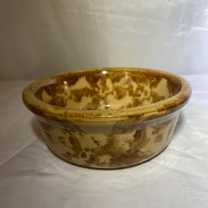 Puede incluir: Un cuenco redondo de cerámica con un patrón moteado marrón y beige. El cuenco tiene un borde ligeramente ensanchado y un acabado brillante. El interior y el exterior del cuenco están decorados con el mismo diseño moteado.