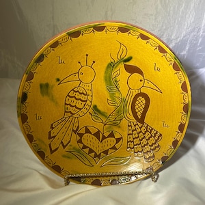 May include: A yellow ceramic plate with a brown rim and a green and brown design of two birds with a heart in the center. The plate has a brown stand.