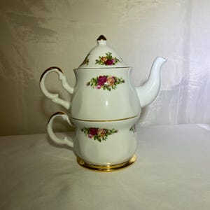 Könnte beinhalten: Weißes Porzellan-Tee-für-eins-Set mit einer Teekanne und einer Tasse, die aufeinander gestapelt sind. Das Set hat ein florales Rosenmuster in Rosa, Rot und Gelb mit goldenen Verzierungen. Die Teekanne hat einen Deckel und einen gebogenen Griff.
