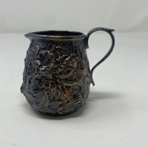 May include: A small, antique silver creamer with a detailed floral pattern. The creamer has a curved handle and a spout for easy pouring. The surface is textured with embossed flowers and leaves, giving it a vintage look.
