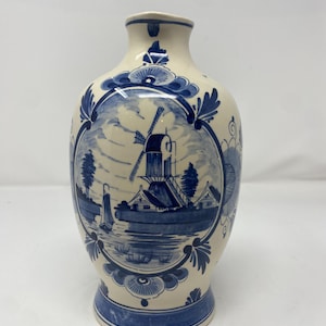 May include: Cream ceramic vase with a blue windmill and landscape design. The vase has a flared base and a narrow neck, with blue floral accents. The artwork depicts a windmill, houses, and a sailboat.
