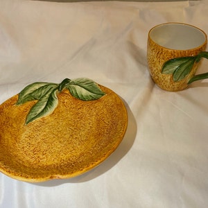 May include: A yellow ceramic plate shaped like a lemon with green leaves. A matching yellow ceramic mug with a green leaf design.
