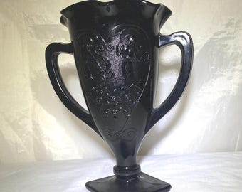 Peut inclure: Vase en verre noir avec deux anses et une base carrée. Le vase a un bord festonné et est décoré de motifs floraux et en forme de cœur en relief. Le vase mesure environ 20 cm de haut.